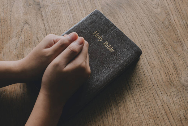 Egy nő Bibliával imádkozik, a kezei össze vannak kapaszkodva a Bibliáján a fa asztalon.. - Fotó, kép