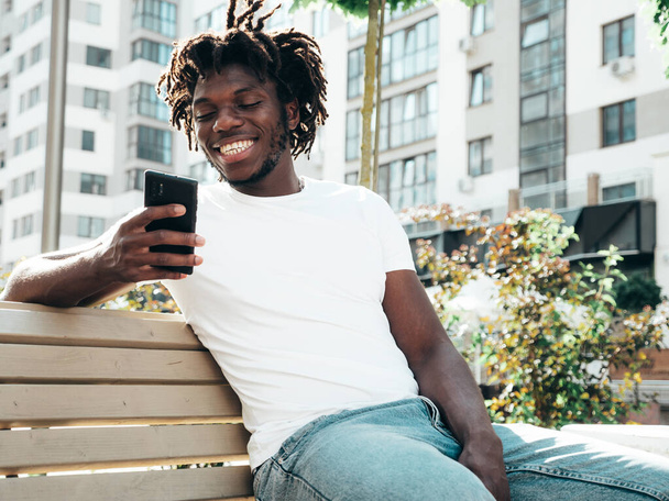 Handsome hipster model.Unshaven African man dressed in white summer t-shirt.Fashion male with dreadlocks hairstyle sitting at the bench in the street.Looking at smartphone screen, using cellphone apps - Foto, Imagen