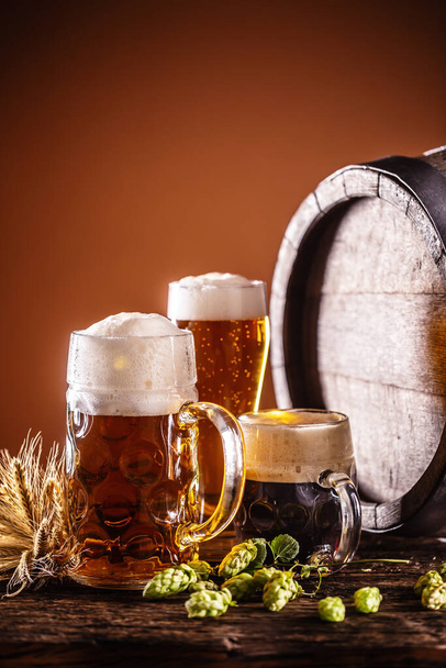 A liter glass full of draft beer next to it two smaller beers in front of a wooden barrel as a decoration of barley and hops. - Zdjęcie, obraz
