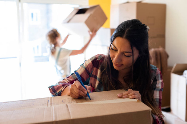 Mladá brunetka běloška píše etiketu na kartonovou krabici pro stěhování domů. Rozumím. Koncept přemísťování. - Fotografie, Obrázek