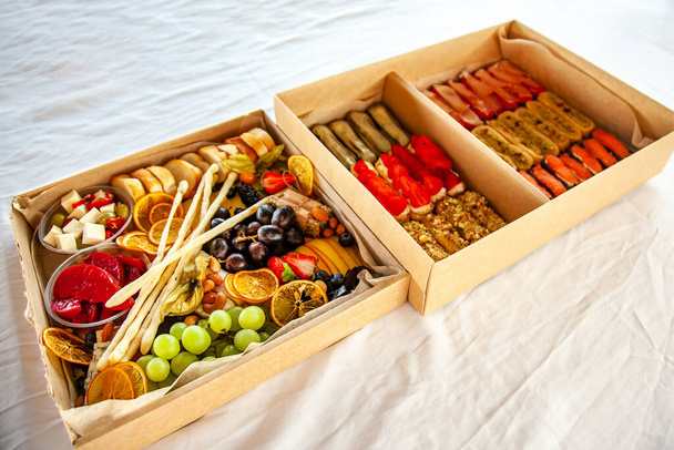 Delicious wine snacks in a cardboard box for a buffet for a party: cheeses, sausages, cold cuts, fruit, baguette, Grissini - Photo, Image