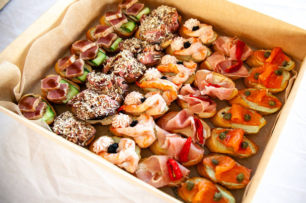 Box with small sandwiches, bruschetta with cold cuts, cheese and seafood. Gourmet snack food in cardboard box for catering buffet for party. - Fotó, kép