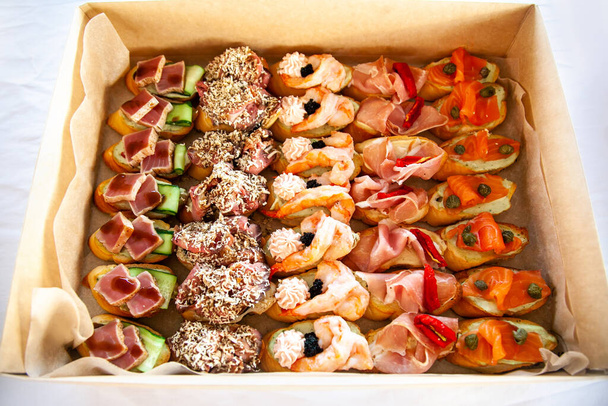 Box with small sandwiches, bruschetta with cold cuts, cheese and seafood. Gourmet snack food in cardboard box for catering buffet for party. - Foto, Imagen