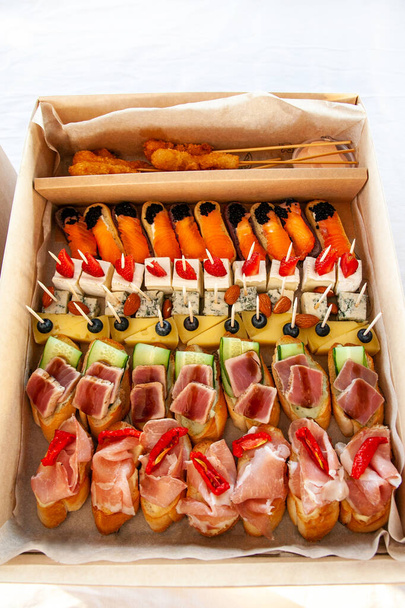 Snack food in cardboard box with small sandwiches, eclairs, bruschetta with cold cuts, cheese and seafood for catering buffet for party. - Photo, Image