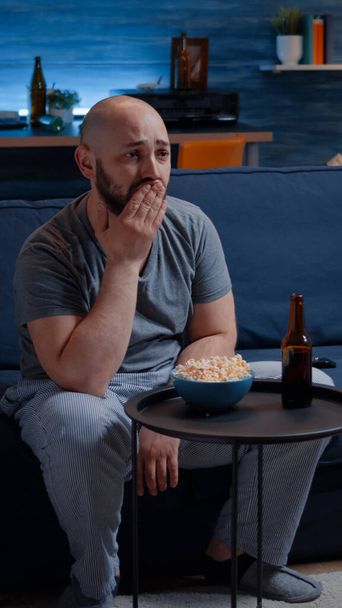 Zaostřený muž se dívá na dramatický film, pláče sedí na gauči a jí popcorn - Fotografie, Obrázek