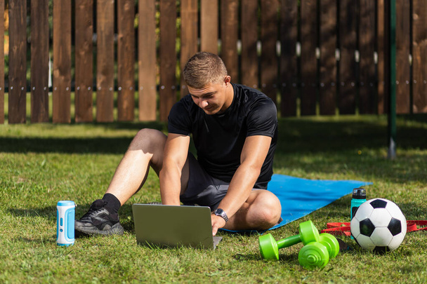 Outdoor portret van de jongeman in een sportuniform rust, strekt zich uit op het gazon, kijkt een film en studeert vanaf een laptop, een sociaal netwerk in de tuin - Foto, afbeelding