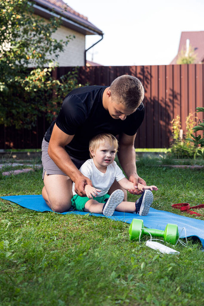 Mladý táta atlet se svým malým veselým synem jde do sportu, protahuje se na rohožce v teplý den v zahradě u domu - Fotografie, Obrázek
