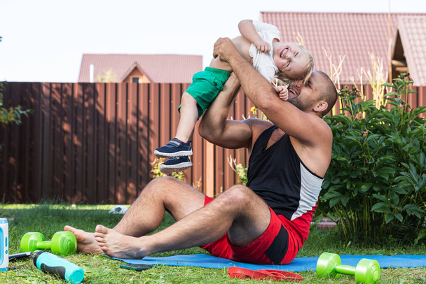 Молодой папа спортсмен со своим маленьким веселым сыном занимается спортом, растягиваясь на коврике в теплый день в саду рядом с домом - Фото, изображение