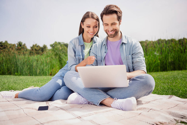 Photo of smiling lovely couple sit outside in park husband browsing in laptop wife point finger buy online shopping sale discount - Photo, image