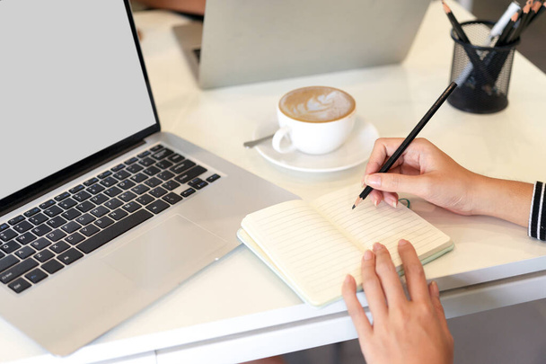 L'immagine ritagliata di donna prende appunti sul notebook accanto al modello di computer portatile e alla tazza di caffè in ufficio - Foto, immagini