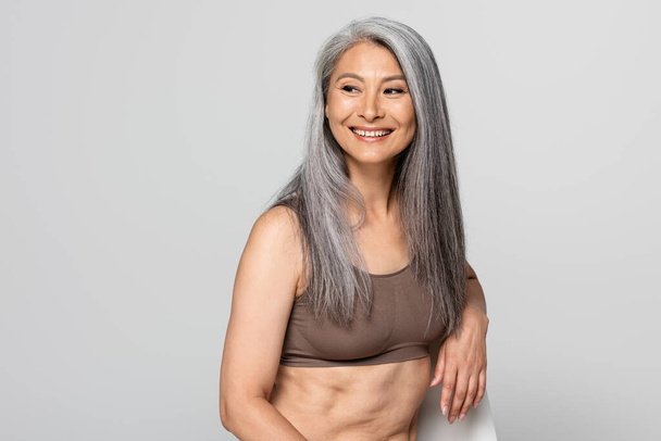Positive asian woman in bra looking away on chair isolated on grey - Foto, immagini