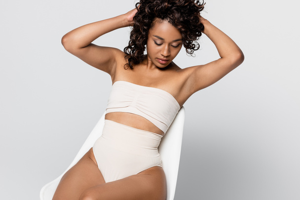 African american woman in underwear adjusting hair on chair isolated on grey - Foto, afbeelding