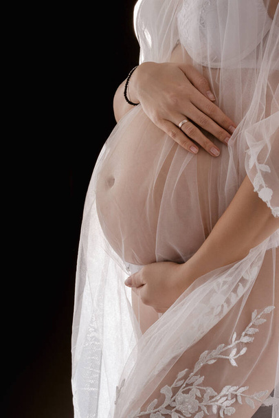 sesión de fotos romántica y de embarazo, mujer embarazada joven en vestido blanco transparente sobre fondo oscuro, luz de estudio, maternidad y concepto de expectativa de bebé - Foto, imagen