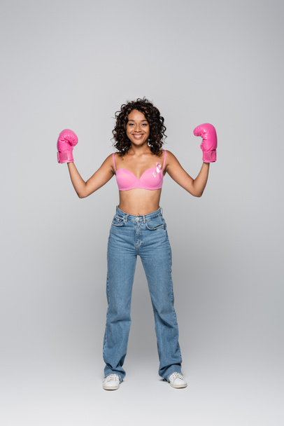 Cheerful african american woman with pink ribbon of breast cancer awareness and boxing gloves on grey background  - Фото, зображення
