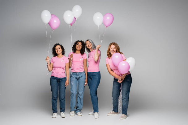 Lächelnde multiethnische Frauen mit rosa Schleifen und Luftballons auf grauem Hintergrund  - Foto, Bild