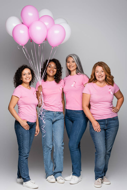 Femmes souriantes multiethniques avec des rubans de sensibilisation au cancer du sein tenant des ballons sur fond gris  - Photo, image