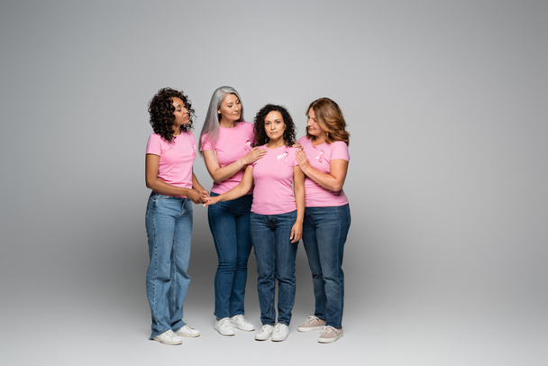 Multiethnic women hugging friend with pink ribbon on t-shirt on grey background  - Foto, imagen