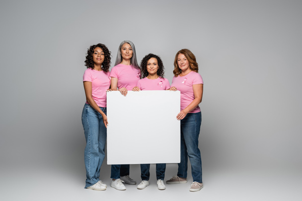 Mujeres multiétnicas con cintas de concienciación sobre el cáncer de mama sosteniendo pancarta sobre fondo gris  - Foto, imagen