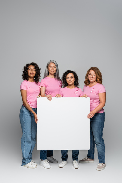Multikulturelle Frauen in rosafarbenen T-Shirts mit Bändern halten leere Plakate auf grauem Hintergrund  - Foto, Bild