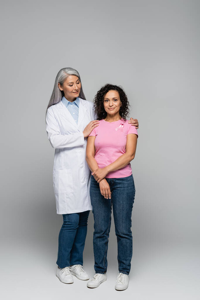 Χαμογελαστή Ασιάτισσα γιατρός αγκαλιάζει Αφροαμερικανή γυναίκα με κορδέλα ευαισθητοποίησης για τον καρκίνο του μαστού σε γκρι φόντο  - Φωτογραφία, εικόνα