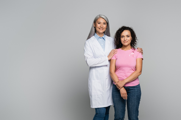 Sorridente medico asiatico in camice bianco che abbraccia la paziente afro-americana con nastro di consapevolezza del cancro al seno isolato su grigio - Foto, immagini