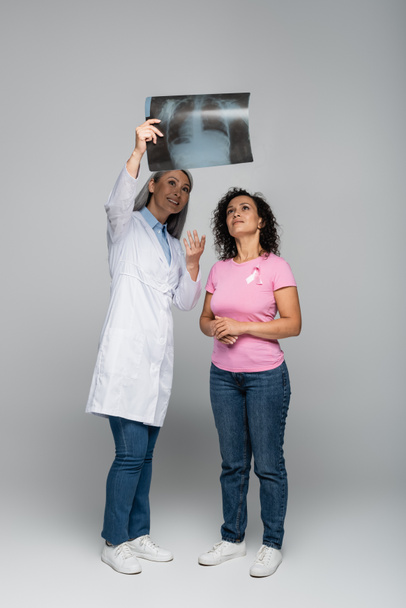 Smiling asian doctor pointing at fluorography near african american woman with ribbon of breast cancer awareness isolated on grey - Photo, Image