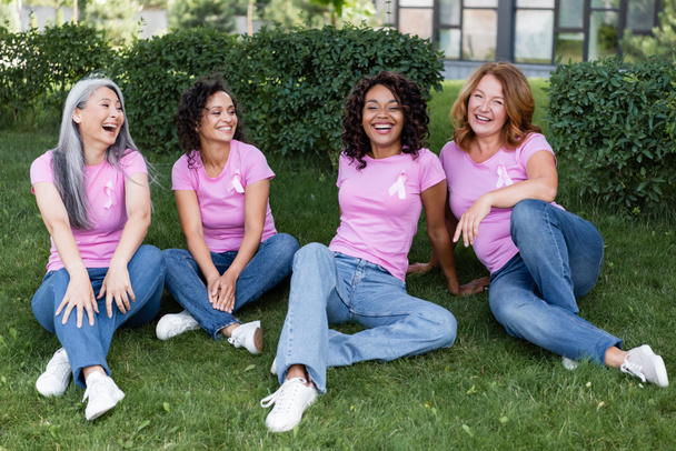 Usmívající se multietnické ženy v růžových tričkách se stuhami sedícími na trávě  - Fotografie, Obrázek