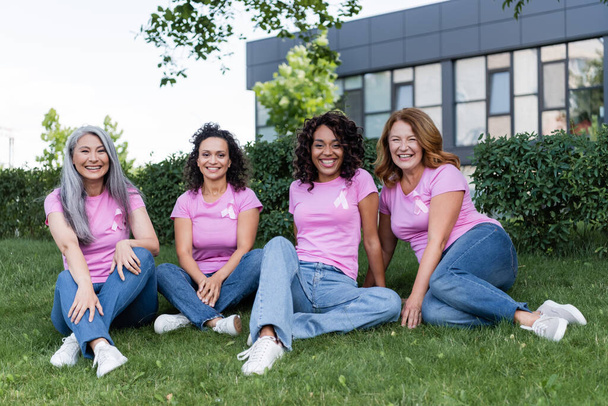 Щасливі міжрасові жінки з рожевими стрічками на футболках, що сидять на траві
  - Фото, зображення