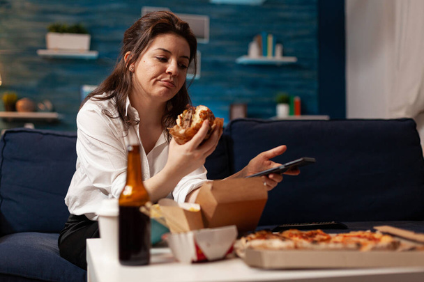 Καυκάσια γυναίκα κρατώντας tastu burgery στα χέρια περιήγηση στα μέσα κοινωνικής δικτύωσης χρησιμοποιώντας smartphone - Φωτογραφία, εικόνα