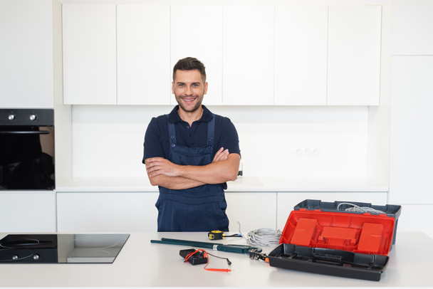 réparateur souriant debout avec les bras croisés près des outils et des fils dans la cuisine - Photo, image