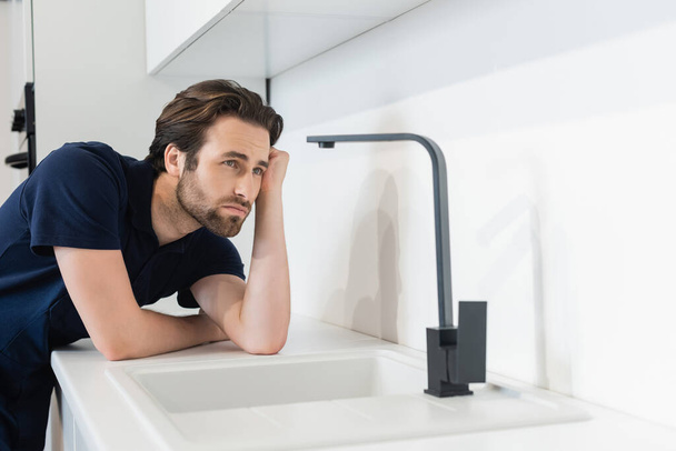 young plumber looking at faucet in modern kitchen - Foto, Imagem