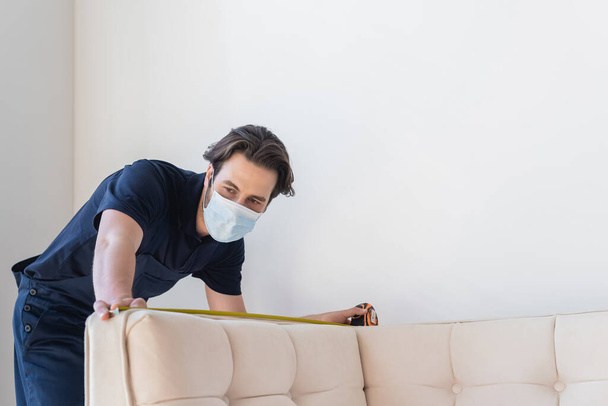 young workmen in medical mask measuring white sofa in apartment  - Фото, изображение