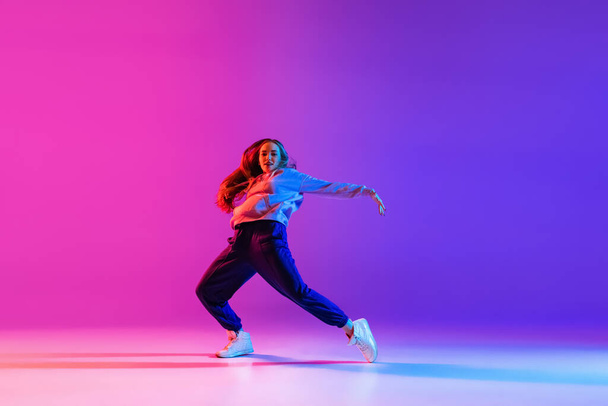Young attractive girl, female hip-hop dancer isolated on gradient pink purple neon studio background - Zdjęcie, obraz