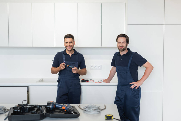 smiling handymen looking at camera near tools on kitchen table - Photo, Image