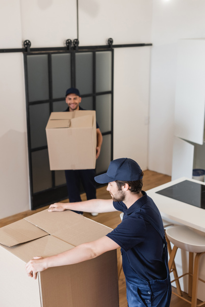 blurred mover holding carton box near colleague in kitchen - Foto, afbeelding