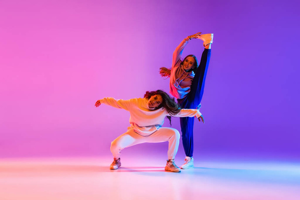Δύο όμορφα νεαρά κορίτσια που χορεύουν χιπ-χοπ σε διαβαθμίσεις ροζ μωβ νέον φόντο στούντιο - Φωτογραφία, εικόνα