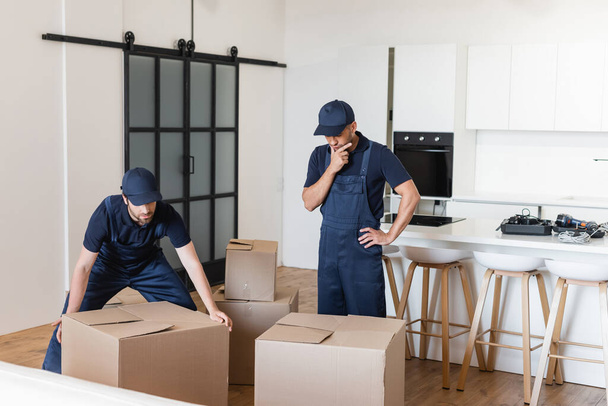 thoughtful worker standing with hand on hip near colleague and carton boxes in kitchen - Photo, Image