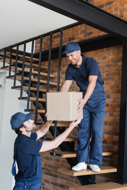 workman giving carton box to smiling mover standing on stairs in apartment - Photo, Image