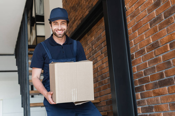 delivery man smiling at camera while walking downstairs with carton package - Photo, Image
