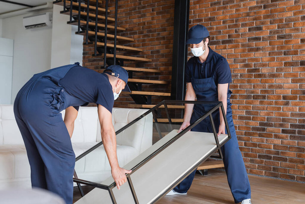 movers in uniform and medical masks lifting glass table near stairs - Photo, Image