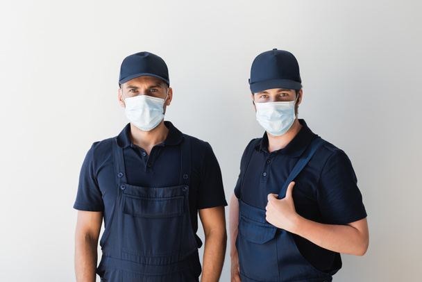workmen in overalls, caps and medical masks looking at camera near white wall - Photo, Image