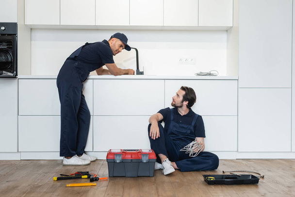plumber looking at workman sitting on kitchen floor near toolbox and tools - Foto, Imagem