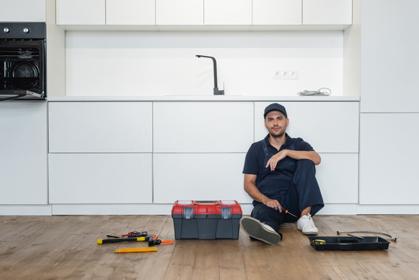 разнорабочий в комбинезоне, сидящий на полу на кухне рядом с инструментами и инструментами - Фото, изображение