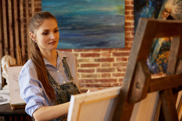 Young beautiful female artist painting on canvas using oil paintings and art brush. Painter creating artwork in art studio. Relaxation, leisure, hobby, stress management - Photo, Image