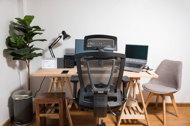 Configuración de escritorio de madera con ordenador portátil, pantalla, ratón, lámpara, silla ergonómica y árbol artificial en el lugar de trabajo en el dormitorio - Foto, imagen