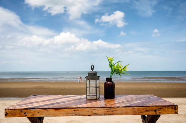 Houten eettafel met lantaarn en bloem in glas op het strand en blauwe lucht in de zomer - Foto, afbeelding