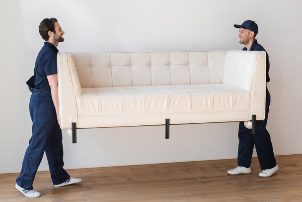 πλαϊνή άποψη των χαμογελαστών εργαζομένων άρση λευκό καναπέ στο διαμέρισμα - Φωτογραφία, εικόνα