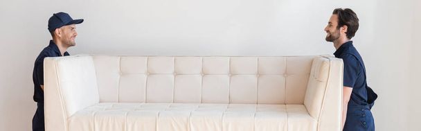 vista lateral de movers sorridentes em uniforme carregando sofá branco, banner - Foto, Imagem
