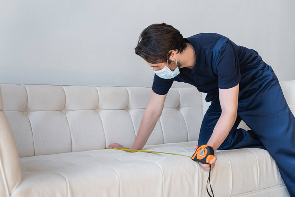 young handyman in medical mask measuring white couch - Foto, Bild