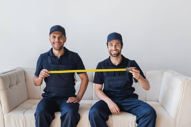 cheerful movers sitting on sofa and holding measuring tape - Photo, Image